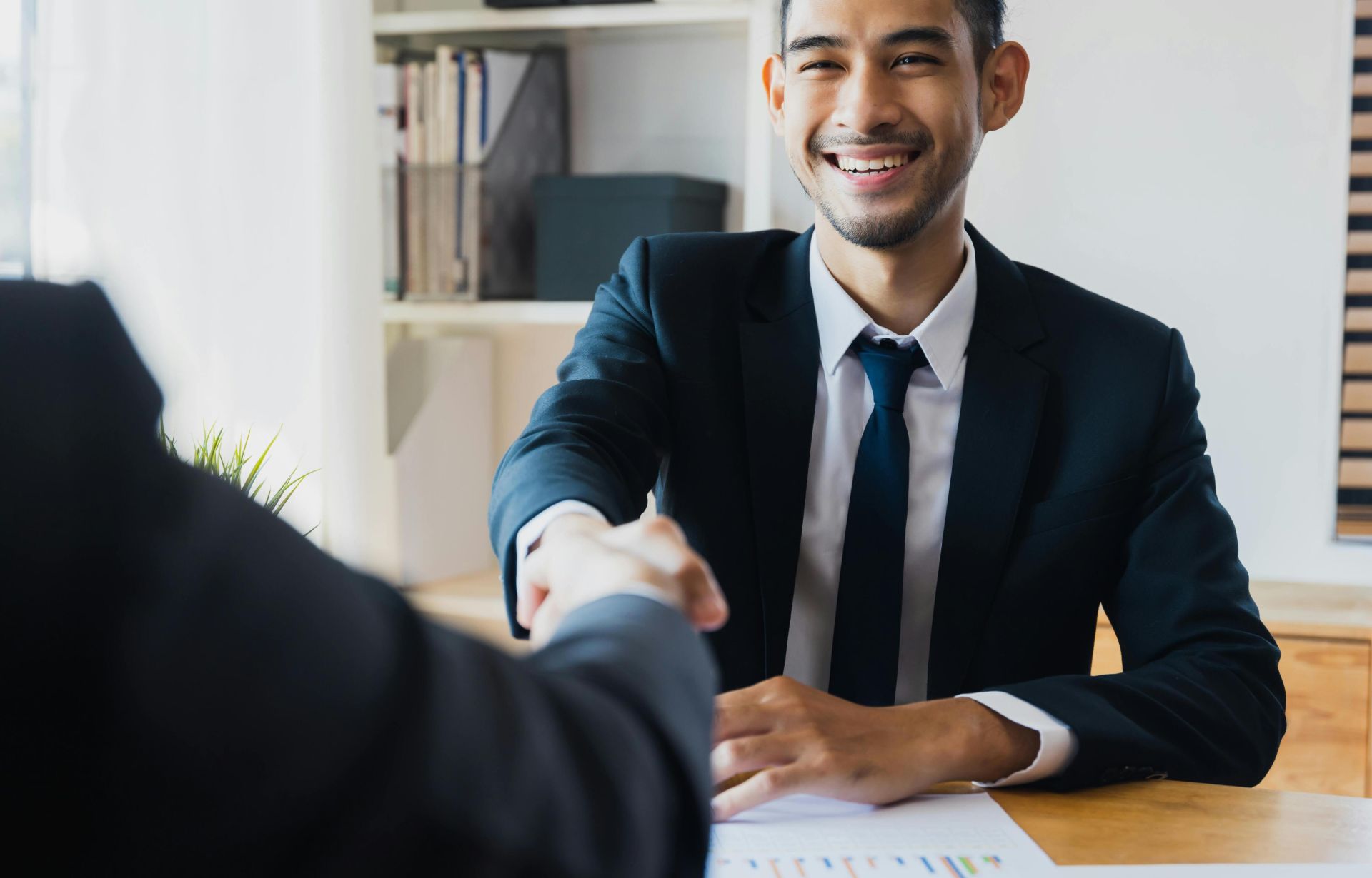 Confident businessman in suit shaking hands at office desk, symbolizing successful partnership.