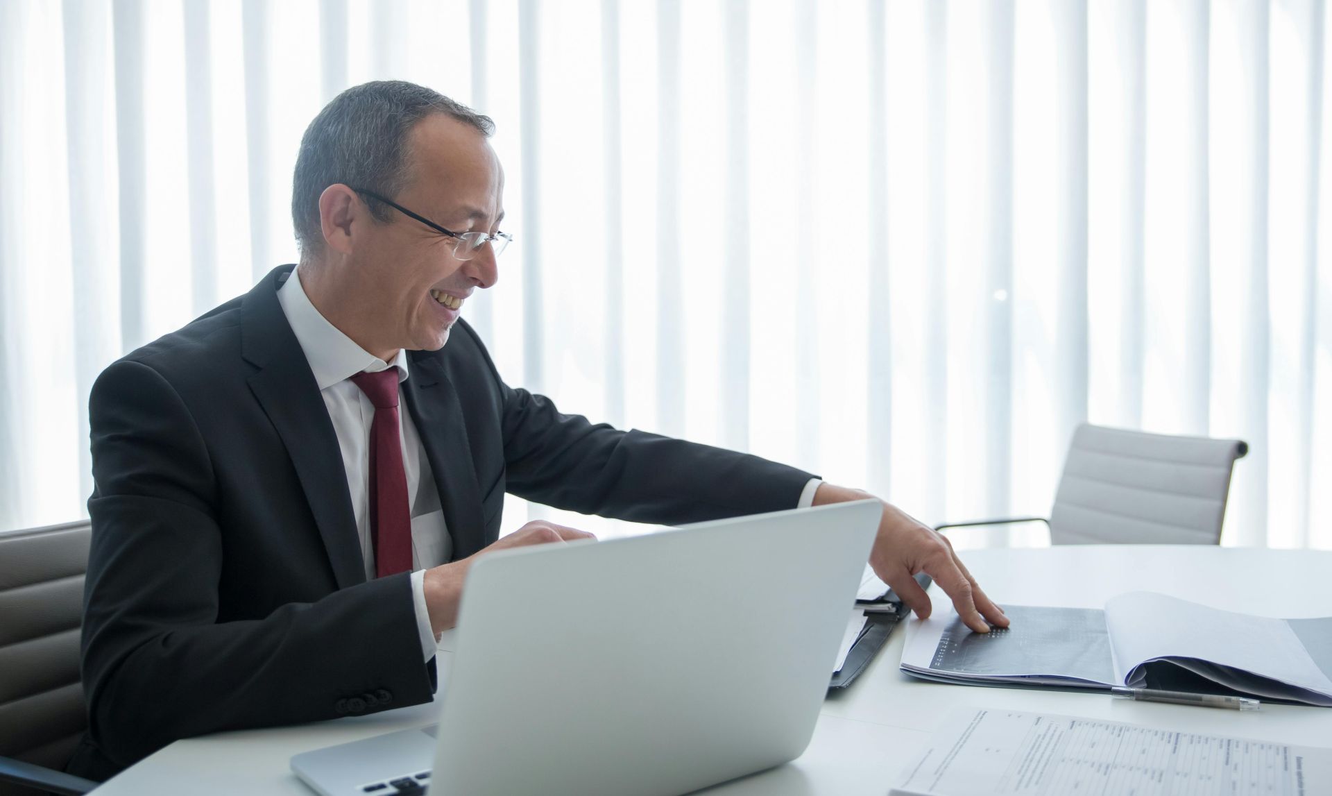 Elderly businessman in a suit working on a laptop and reviewing documents in a bright office setting.